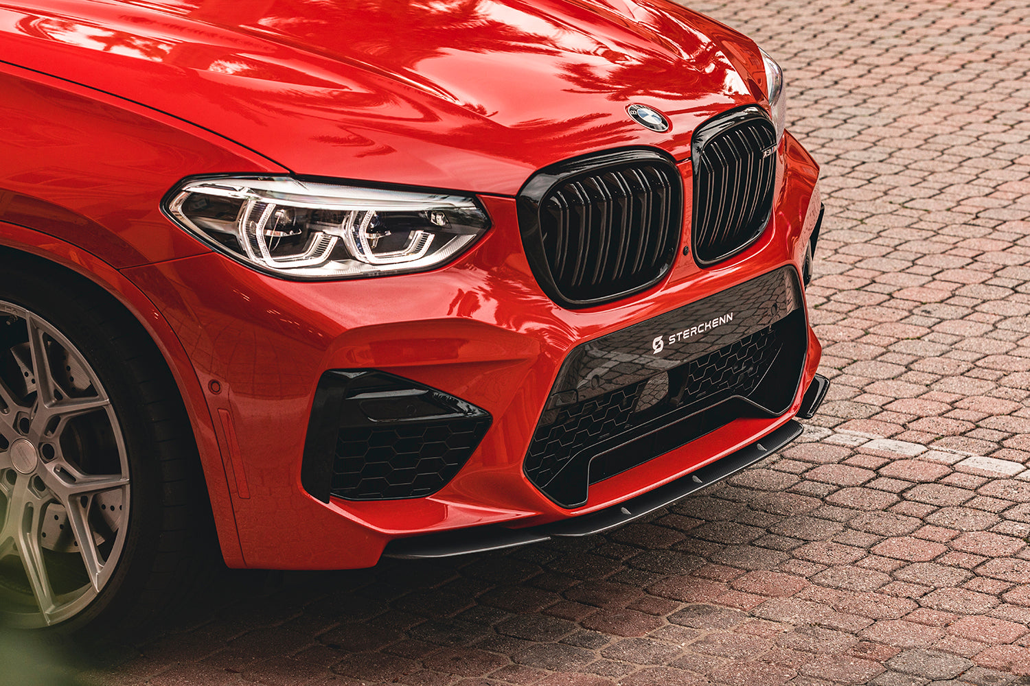 The bumper of a red BMW X3M (F97) against a background of paving stones
